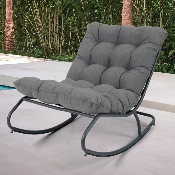 Fotel bujany AGAVA Dark Grey w aranżacji ogrodowej, biała posadzka, drzewa i krzewy w tle Fotel bujany AGAVA Dark Grey w aranżacji ogrodowej, biała posadzka, drzewa i krzewy w tle