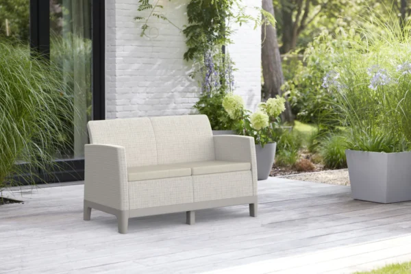 Sofa ogrodowa Keter Scandi Forma 2 w kolorze Pebble Grey-Soft Beige Oat stoi na drewnianym tarasie pośród zieleni