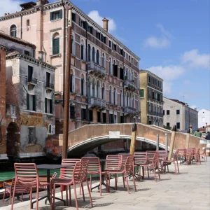 Różowe krzesła Nardi DOGA BISTROT Bianco ustawione przy stolikach na kamiennym deptaku z widokiem na kanał i budynki Różowe krzesła Nardi DOGA BISTROT Bianco ustawione przy stolikach na kamiennym deptaku z widokiem na kanał i budynki