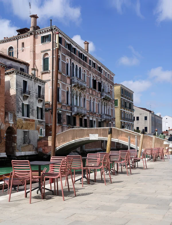 Różowe krzesła Nardi DOGA BISTROT Bianco ustawione przy stolikach na kamiennym deptaku z widokiem na kanał i budynki Różowe krzesła Nardi DOGA BISTROT Bianco ustawione przy stolikach na kamiennym deptaku z widokiem na kanał i budynki