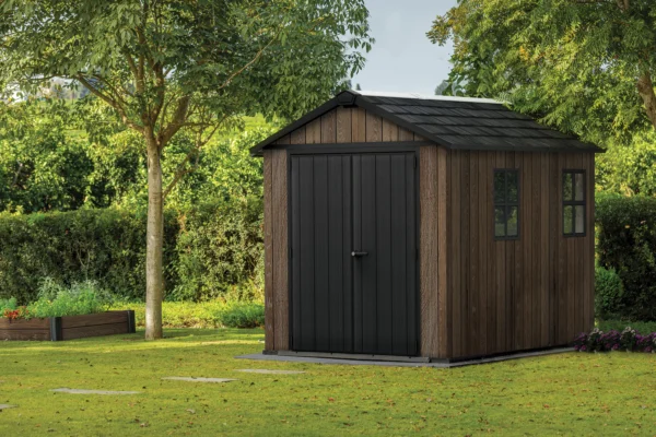 Domek narzędziowy Keter DECO APEX 759/975 orzechowy z czarnymi drzwiami i oknami w otoczeniu zieleni Domek narzędziowy Keter DECO APEX 759/975 orzechowy z czarnymi drzwiami i oknami w otoczeniu zieleni
