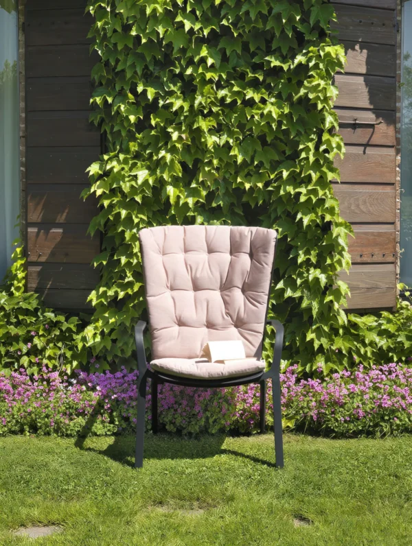 Poduszka na fotel FOLIO COMFORT Rosa Quarzo Sunbre – Nardi na fotelu w ogrodzie na tle kwiatów i drewnianej elewacji