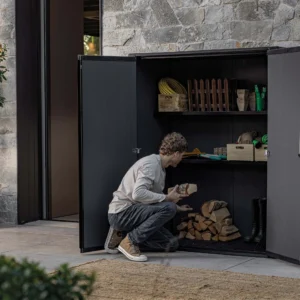 Mężczyzna układający drewno w otwartym brązowym schowku ogrodowym Deco Vertical 1400L Venetian Brown Keter na tarasie