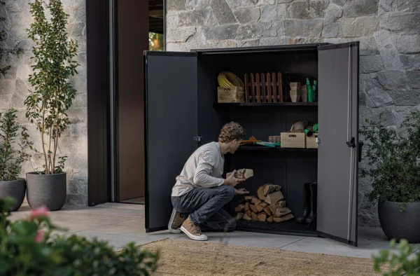 Mężczyzna układający drewno w otwartym brązowym schowku ogrodowym Deco Vertical 1400L Venetian Brown Keter na tarasie