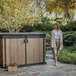 Szafa wielofunkcyjna Deco Horizontal 2020L Venetian Brown Keter w ogrodowej aranżacji z kobietą spacerującą po ścieżce