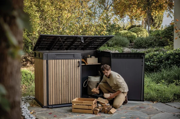 Mężczyzna układający drewno obok szafy wielofunkcyjnej Deco Horizontal 2020L Venetian Brown Keter z otwartymi drzwiami i wiekiem